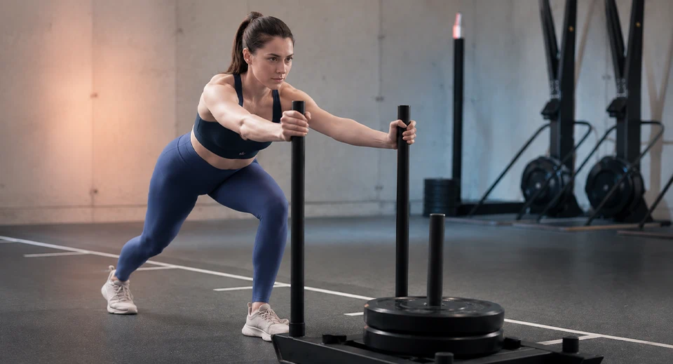 Woman driving a weighted sled across turf, a core HYROX training movement